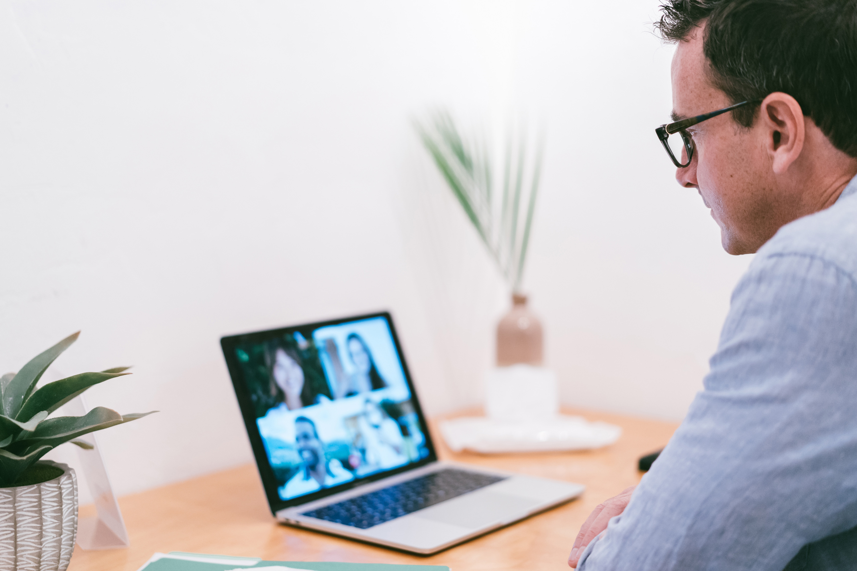 A man wearing glasses engaged in a video conference on a laptop at a neatly arranged wooden desk with a plant and decorative elements.